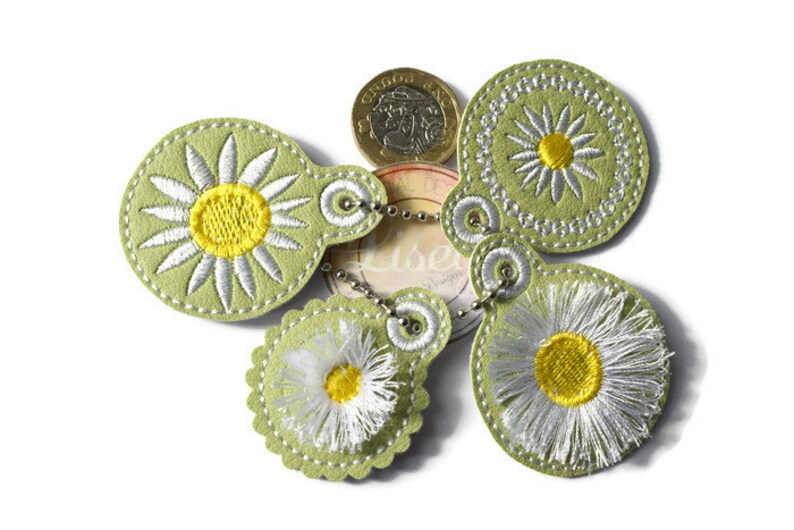 May include: Five green felt keyrings with daisy embroidery. Each keyring has a different style of daisy, with white petals and a yellow centre. The keyrings are connected by a silver chain.