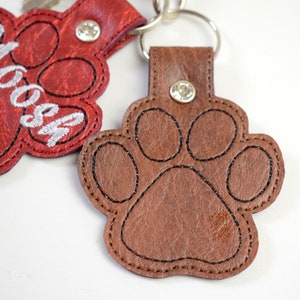 May include: Two leather paw print keyrings. One is red with the name "Moosh" embroidered in white. The other is brown.
