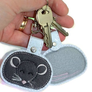 May include: A white and gray felt keychain with a cute embroidered mouse face on one side and a plain gray side on the other. The keychain is attached to a silver key ring.