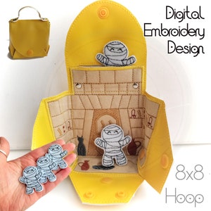 May include: A yellow felt pouch with a flap opening, decorated with mummy characters. The pouch is open to reveal a felt interior with a mummy character and other details. The pouch is designed for an 8x8 hoop embroidery project.  Digital Embroidery Design.