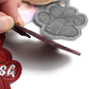 May include: Red leather paw print keyring with the name "Boo" embroidered in white thread. The keyring has a purple leather strap and a silver metal ring.