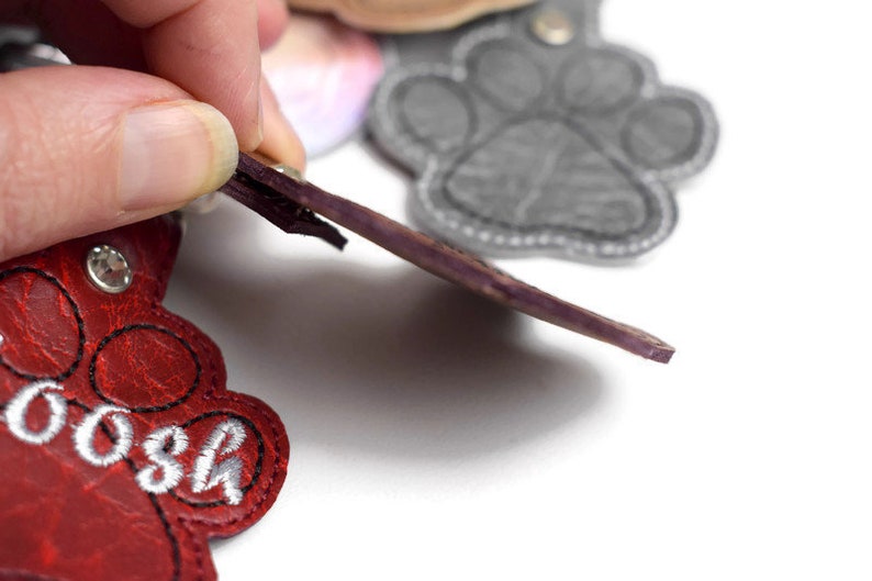 May include: Red leather paw print keyring with the name "Boo" embroidered in white thread. The keyring has a purple leather strap and a silver metal ring.