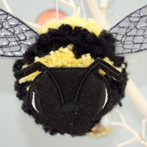 May include: A black and yellow fuzzy bee ornament with black felt wings and a black felt face with white embroidered eyes.