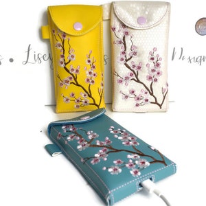 Puede incluir: Tres fundas para teléfono en amarillo, crema y turquesa, cada una con una rama de flores de cerezo bordada. Las fundas tienen un cierre de solapa con un broche y un lazo. La funda turquesa tiene un cable de carga.