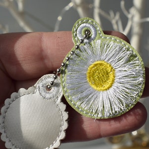 May include: A white leather keychain with a green daisy design and a silver chain. The daisy has a yellow centre and white petals.