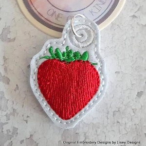 May include: A white felt strawberry charm with a red embroidered strawberry and green embroidered leaves. The charm has a silver loop for attaching to a keychain or bag.