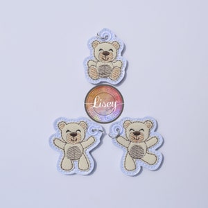 Könnte beinhalten: Drei gestickte Teddybär-Schlüsselanhänger mit braunem Fell, weißem Gesicht und schwarzen Augen. Die Schlüsselanhänger befinden sich auf einem hellblauen Hintergrund.