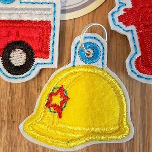 May include: Three embroidered felt keychains with a firefighter theme. The keychains are a red fire engine, a yellow firefighter helmet with a red star, and a red fire hydrant.