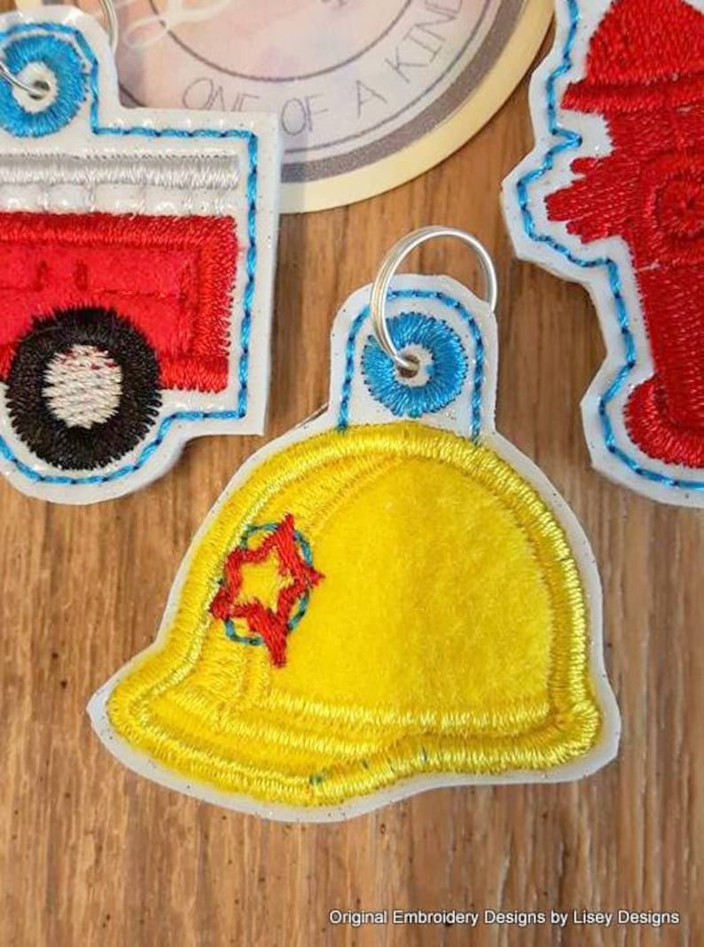 May include: Three embroidered felt keychains with a firefighter theme. The keychains are a red fire engine, a yellow firefighter helmet with a red star, and a red fire hydrant.