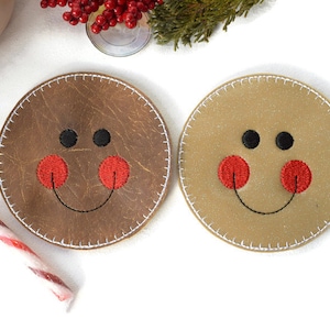 May include: Two round brown and beige coasters with a stitched edge. Each coaster has an embroidered smiley face with black eyes, a red nose, and a black stitched smile.