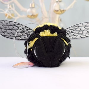 May include: A black and yellow bumble bee ornament with embroidered wings. The bee is made of yarn and has a fuzzy texture.
