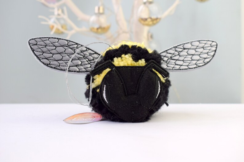 May include: A black and yellow bumble bee ornament with embroidered wings. The bee is made of yarn and has a fuzzy texture.