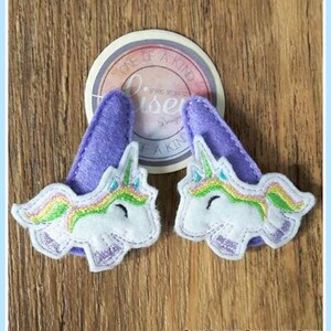 May include: Two purple felt unicorn hair clips with rainbow manes and white faces. The clips are decorated with a rainbow and a white unicorn face. The text "One of a Kind" is visible on a white circle behind the clips.
