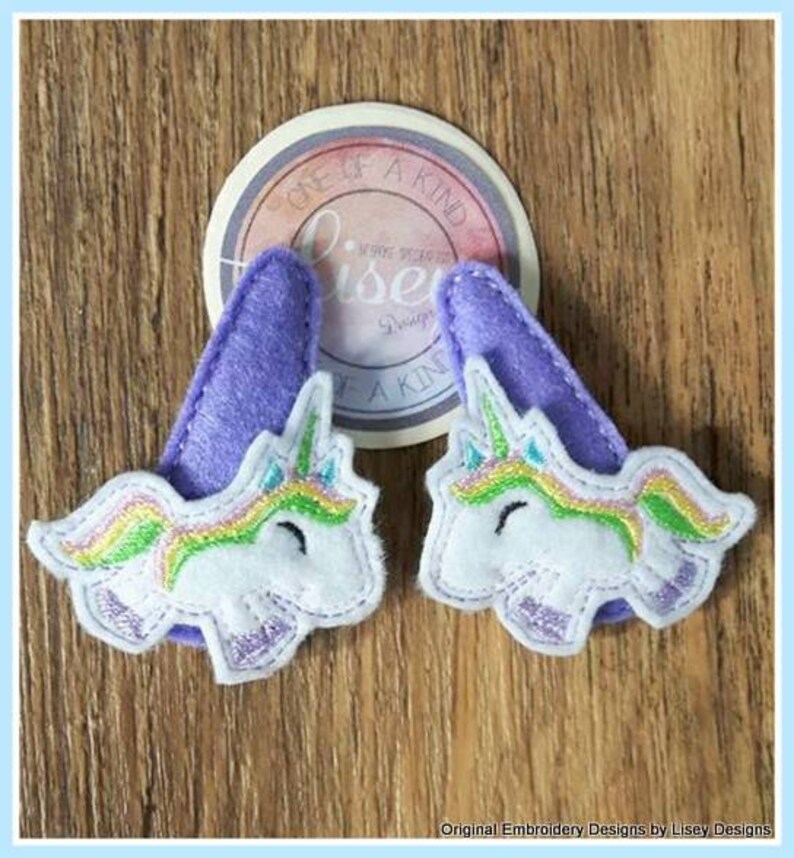 May include: Two purple felt unicorn hair clips with rainbow manes and white faces. The clips are decorated with a rainbow and a white unicorn face. The text "One of a Kind" is visible on a white circle behind the clips.