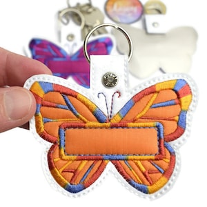 Puede incluir: Llavero de mariposa bordado en naranja, rojo, azul y amarillo con una etiqueta de nombre naranja en blanco. El llavero está unido a un lazo de tela blanco con un anillo de metal plateado.
