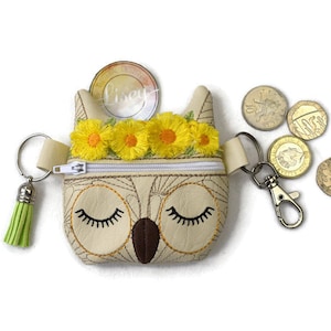 May include: A small beige owl-shaped zippered pouch with a green tassel and a silver key chain. The pouch has embroidered yellow flowers and closed eyes. The pouch is filled with British coins.