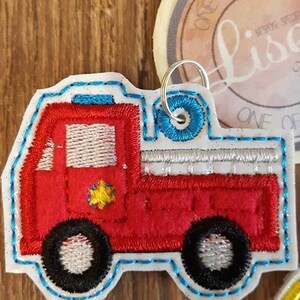 May include: Red and white embroidered fire truck applique with a blue outline. The truck has a yellow star on the side and a silver ladder on the back. The applique is attached to a silver key ring.