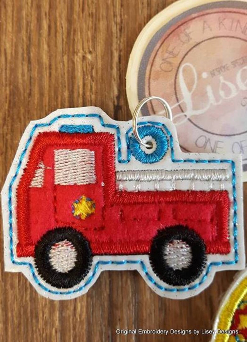 May include: Red and white embroidered fire truck applique with a blue outline. The truck has a yellow star on the side and a silver ladder on the back. The applique is attached to a silver key ring.
