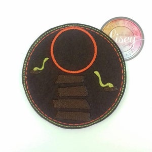 May include: A brown felt circle with an orange circle and brown steps embroidered on it. Two green worms are embroidered on either side of the steps. The circle has a green stitched border.