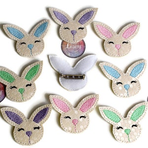 May include: Nine embroidered bunny pins with different colored ears. The bunnies are all beige with a honeycomb pattern and have black eyes and a black nose. The pins are perfect for adding a touch of whimsy to any outfit.