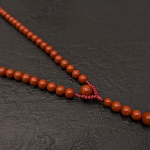 May include: A necklace featuring a series of smooth, round, terracotta-colored beads strung on a matching dark red cord. The design includes a central knot detail that separates the main strand from a short, dangling section of smaller beads.