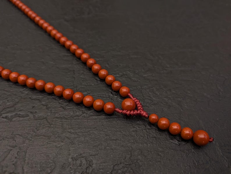 May include: A necklace featuring a series of smooth, round, terracotta-colored beads strung on a matching dark red cord. The design includes a central knot detail that separates the main strand from a short, dangling section of smaller beads.