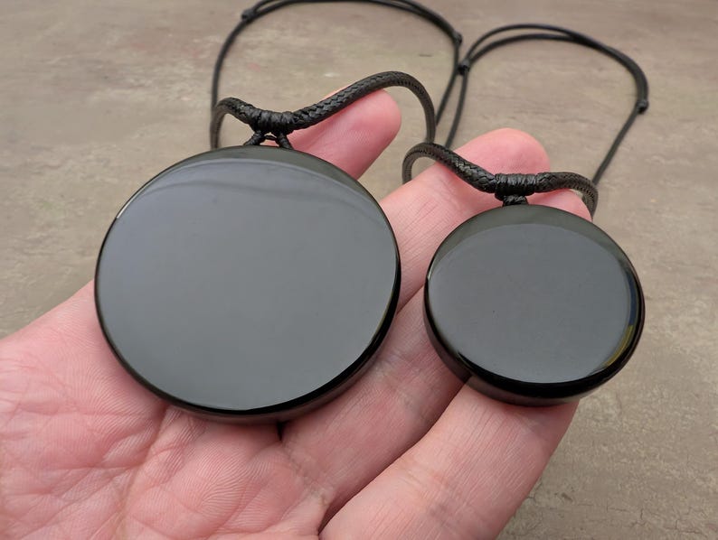 K&ouml;nnte beinhalten: Zwei schwarze Obsidian-Anh&auml;nger an schwarzen Schn&uuml;ren. Der gr&ouml;&szlig;ere Anh&auml;nger ist rund und hat einen Durchmesser von etwa 5 cm. Der kleinere Anh&auml;nger ist ebenfalls rund und hat einen Durchmesser von etwa 2,5 cm.