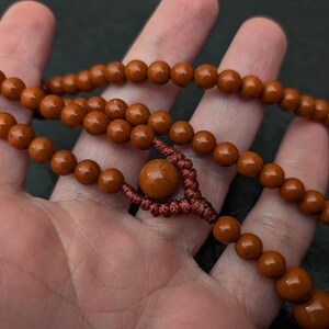 May include: A strand of smooth, round, reddish-brown beads strung together with a dark brown cord. The necklace features a central, larger bead secured by a decorative, intricate knot pattern, with smaller beads continuing along the cord.