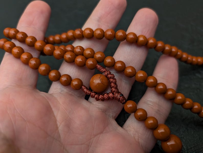 May include: A strand of smooth, round, reddish-brown beads strung together with a dark brown cord. The necklace features a central, larger bead secured by a decorative, intricate knot pattern, with smaller beads continuing along the cord.