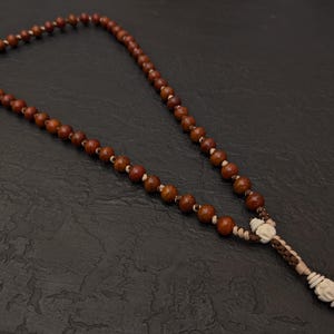 May include: A brown beaded necklace with a tassel. The necklace features round, wooden beads and a decorative tassel with white and tan accents. The beads are strung together with a thin, light-colored string.