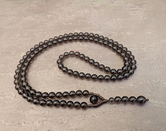 May include: A necklace composed of translucent, dark gray smoky quartz beads strung on a dark brown cord. The design features a continuous loop of beads that transitions into a single strand ending with a larger bead, secured by brown knotting.