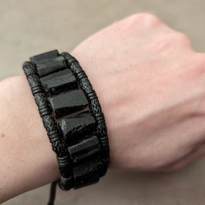 Puede incluir: Un brazalete negro de cuentas con un cordón trenzado. El brazalete está hecho de varias filas de cuentas negras y tiene un cordón trenzado que se ata alrededor de la muñeca.