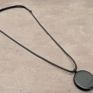 K&ouml;nnte beinhalten: Eine schwarze Halskette mit einem gro&szlig;en, runden, schwarzen Anh&auml;nger. Die Halskette besteht aus einem d&uuml;nnen, schwarzen Band.