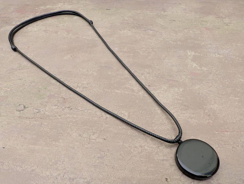 K&ouml;nnte beinhalten: Eine schwarze Halskette mit einem gro&szlig;en, runden, schwarzen Anh&auml;nger. Die Halskette besteht aus einem d&uuml;nnen, schwarzen Band.