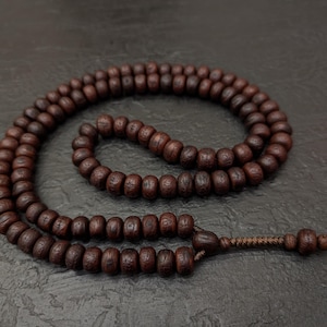 May include: A strand of dark brown wooden prayer beads arranged in a circular loop. Each individual bead features a small, carved circular symbol on its surface. The beads are connected by a brown cord that ends in a decorative knot.