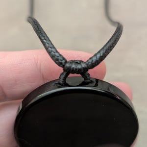 K&ouml;nnte beinhalten: Ein schwarzer Obsidian-Anh&auml;nger mit einer glatten, polierten Oberfl&auml;che, der an einem schwarzen Band h&auml;ngt.