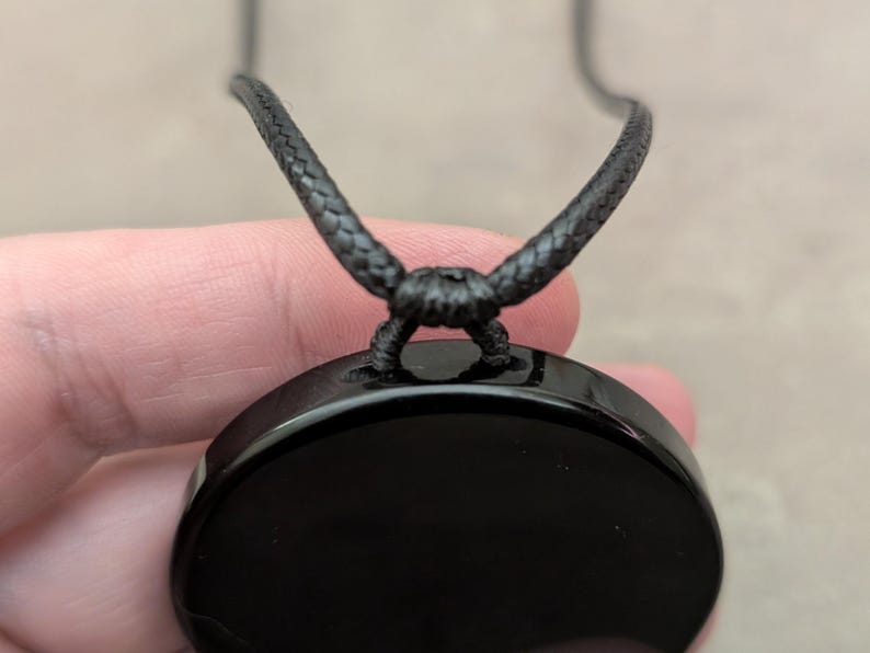 K&ouml;nnte beinhalten: Ein schwarzer Obsidian-Anh&auml;nger mit einer glatten, polierten Oberfl&auml;che, der an einem schwarzen Band h&auml;ngt.
