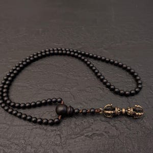 May include: A black beaded necklace with a decorative pendant. The necklace features small, round beads and a detailed, ornate pendant with a bronze finish. The necklace is displayed on a dark surface.