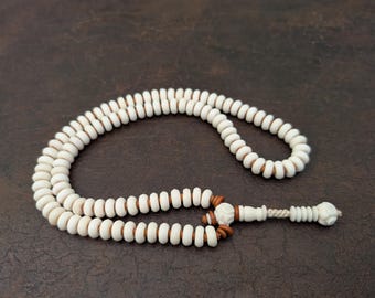 May include: Ivory-colored prayer beads with brown spacers and a decorative tassel. The beads are strung together, forming a loop. The tassel features a carved design and a decorative end piece.
