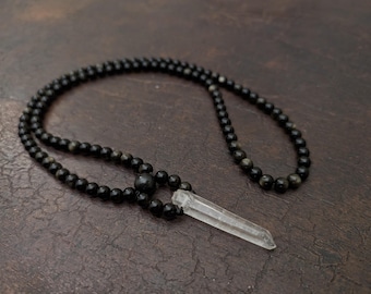 May include: A black beaded necklace with a clear crystal pendant. The necklace features small, round black beads strung together, with a larger black bead at the center. The crystal pendant is a clear, elongated shape.