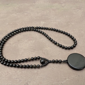 May include: A black beaded necklace with a large, round black pendant. The necklace is made of small, round beads and has a simple design.