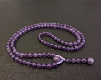 May include: A purple amethyst mala necklace with a tassel. The necklace is made of round beads and is strung on a thin cord.