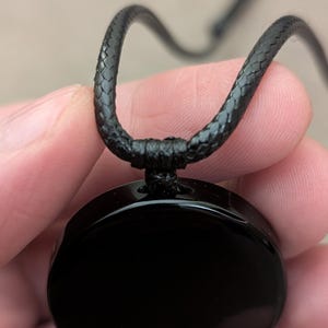 K&ouml;nnte beinhalten: Eine schwarze Halskette mit einem runden, schwarzen Anh&auml;nger. Der Anh&auml;nger ist glatt und gl&auml;nzend. Die Halskette besteht aus einer schwarzen geflochtenen Schnur.