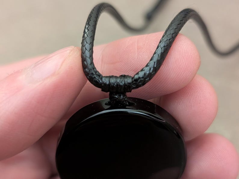K&ouml;nnte beinhalten: Eine schwarze Halskette mit einem runden, schwarzen Anh&auml;nger. Der Anh&auml;nger ist glatt und gl&auml;nzend. Die Halskette besteht aus einer schwarzen geflochtenen Schnur.