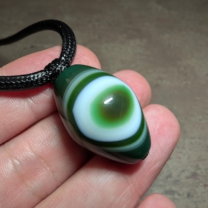 Puede incluir: Un colgante de forma ovalada con un patrón de ojo en capas con anillos concéntricos de color verde oscuro, blanco y un centro marrón profundo. La cuenta de piedra lisa y pulida cuelga de un cordón trenzado negro grueso que pasa por arriba.