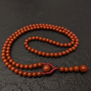 May include: A strand of round, reddish-brown beads arranged in a circular loop. The beads are strung on a matching cord, featuring a decorative knot detail near the end, followed by a series of smaller beads and one larger terminal bead.