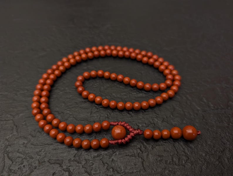 May include: A strand of round, reddish-brown beads arranged in a circular loop. The beads are strung on a matching cord, featuring a decorative knot detail near the end, followed by a series of smaller beads and one larger terminal bead.