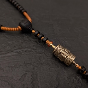 May include: A black beaded necklace with a small, gold-colored prayer wheel pendant. The necklace is strung with orange thread and has a black wooden bead at the top.