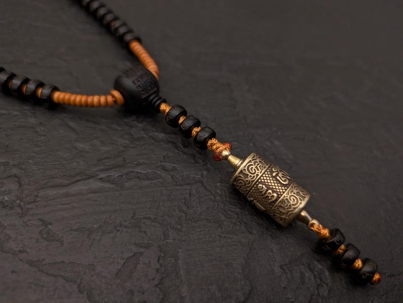 May include: A black beaded necklace with a small, gold-colored prayer wheel pendant. The necklace is strung with orange thread and has a black wooden bead at the top.