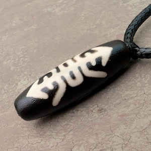 May include: A black pendant with a white design, hanging from a braided black cord. The pendant is oblong with a rounded shape and a white, stylized pattern. The necklace is likely a piece of jewellery.